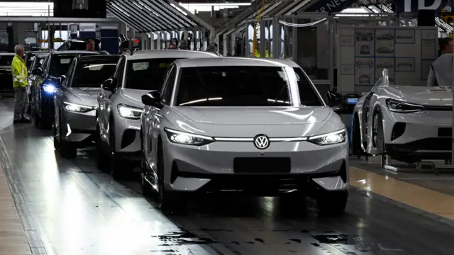 Silvery Volkswagen ID.7 electric vehicles in a line in a darkened factory, with a man in a green jacket standing near one