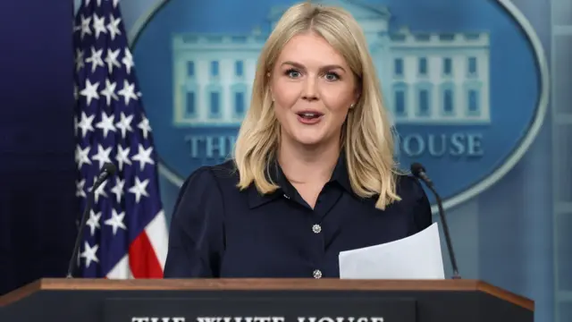 Leavitt in dark blouse at the press podium in front of American flag