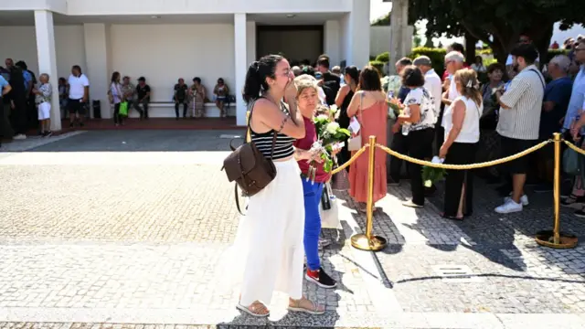 A woman, looking tearful, holds her hand over her mouth near queuing mourners