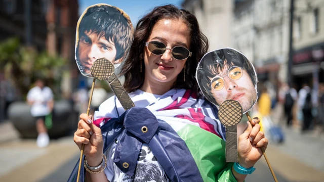 A woman with long dark hair wearing a Welsh flag draped round her shoulders and sunglasses. In each hand she has a wooden stick with a cutout of Liam and Noel Gallagher on each.
