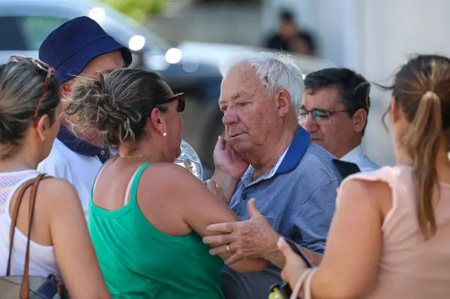 A woman comforting Jota and Silva's grandfather who is surrounded by mourners