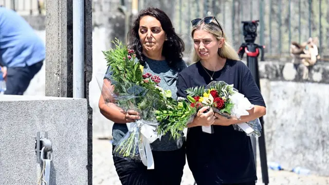 A brown-haired woman (L) and a young blonde woman (R) hold flower tributes as they walk under the sun towards the Chapel of the Resurrection in Gondomar, Portugal