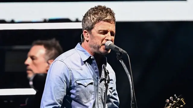 Noel Gallagher with acoustic guitar on stage.