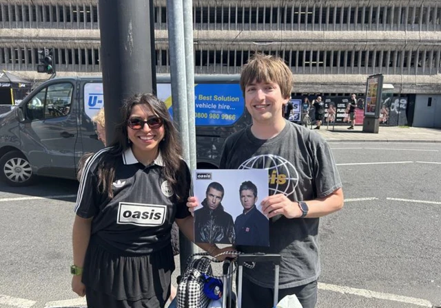 A woman with long dark hair and a man with brown hair, both wearing black and stood on a pavement with a road behind them. The woman wears sunglasses and the man is holding up an oasis album. They are both smiling at the camera.