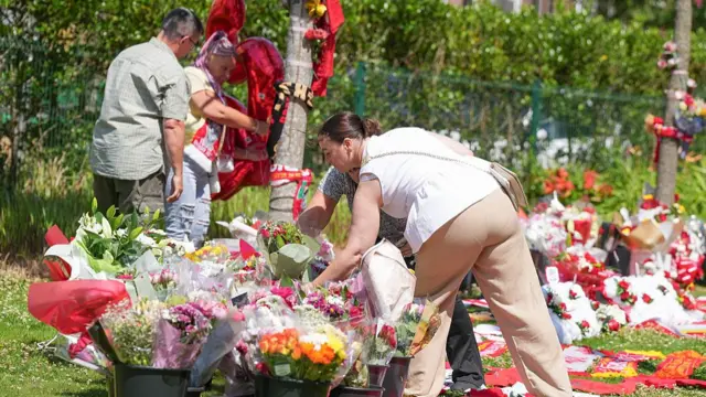 A woman in a white flowy tank top and beige trousers lays flowers outside Anfield Stadium. Several flower tributes and Liverpool FC scarves rest on a grassy patch
