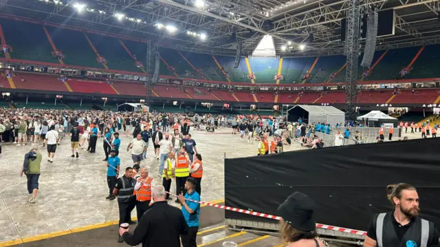 Crowds of people on the floor of a stadium, with stands of red chairs surrounding them.