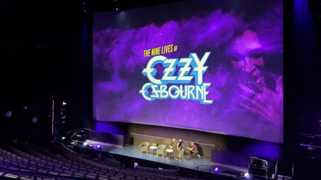 An auditorium that is lit up purple as a result of a large screen that has a purple holding screen. The screen reads "The Nine Lives of Ozzy Osbourne" and a picture of an older's man face is to the right.