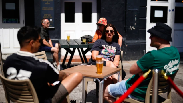 A group of three people sat at a table outside a pub. A woman, facing the camera, is wearing an oasis t shirt and sunglasses and has long brown hair. Shehas apint in front of her. She is facing two men, on is wearing a black t shirt and sunglasses, the other a green t shirt with denim shorts and a black bucket hate.