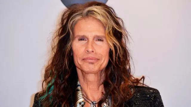 An older man, Steven Tyler, with long, wavy hair, which is mainly auburn but blonde at the front. His lips are pursed and he is wearing a black, sparkly jacket with shoulder pads.