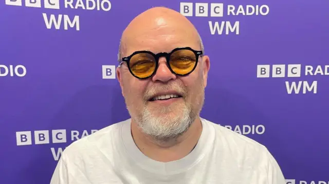 A bald man who is wearing glasses with yellow lenses and a white t-shirt. He is standing in front of a purple screen that reads "BBC Radio WM".