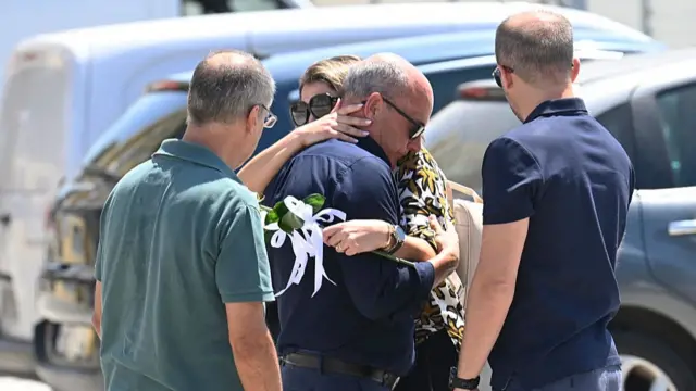 Jota's father hugging a woman holding a flower with white ribbons attached