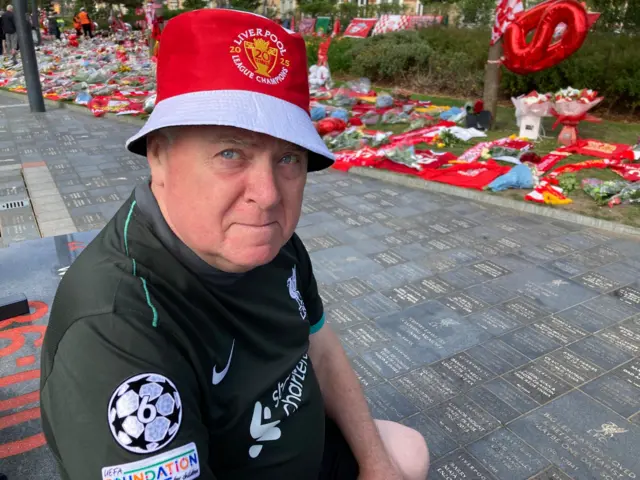 Peter Millea, wearing a red and white Liverpool bucket hat a black replica Liverpool away kit, sits on a bench in front of a sea of red shirts, scarves, flowers and balloons