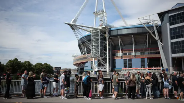 A queue of people on the pavement with the Taff river behind them and the stadium behind that.