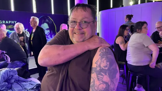 A man with a full sleeve tattoo that is a picture of a man, Ozzy Osbourne, with his mouth open as if he is screaming. The man is sitting down and has short, thin, dark hair. He is wearing glasses and a grey top which he has rolled up to show off his tattoo.