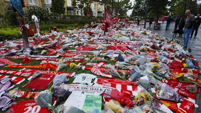 Close up view of tributes to Diogo Jota laid on grassy patch outside Anfield Stadium, people standing to the