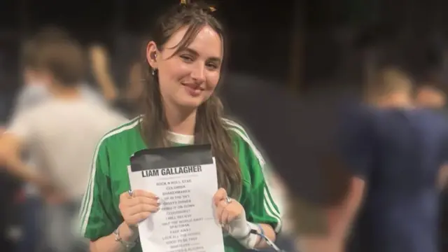 A woman with long brown wearing a green and white Adidas top and holding a Liam Gallagher concert setlist.