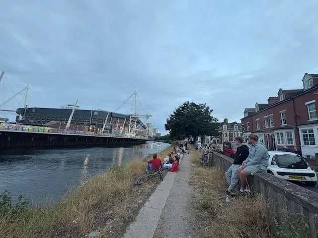 People sat on the walls of the River Taff in Cardiff, they face the Principality stadium and listen on the the Oasis gig.
