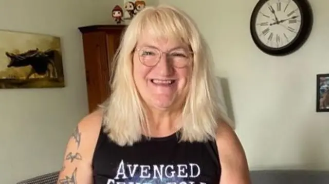 A woman with blonde hair and tattoos on her arms. She is standing a lounge, in front of a cream wall, wearing a black vest top. She is smiling.