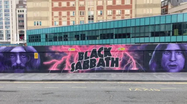 A black, pink and purple wall art mural that reads Black Sabbath in big, bubble writing. Tall buildings are behind the mural