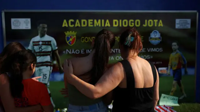 A woman in a black tank top hold her arm around a young girl with a ponytail as they observe tributes to Jota outside the Academia Diogo Jota in Gondomar