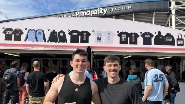 Two boys wearing black t shirts and standing in front of a busy merchandise stand, with the Principality Stadium behind. They both have brown hair, the one on the right has a moustache, and they are both smiling and looking at the camera with an arm round each other.