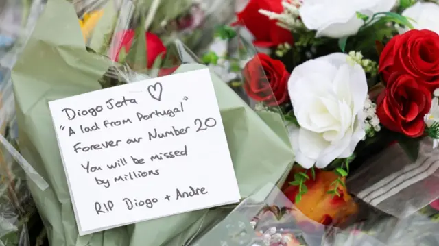 A card reading "Diogo Jota 'a lad from Portugal' Forever our Number 20. You will be missed by millions RIP Diogo+Andre" laying on several bunches of flowers, including white and red roses, outside Anfield
