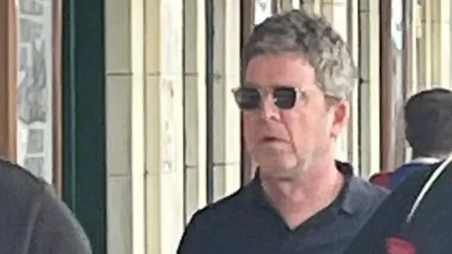 Noel Gallagher on a platform at Cardiff Central Railway Station. He is wearing a black, collared T-shirt and black sunglasses with clear frames. There are three unidentifiable people around him as he walks through a crowd and white brick walls are behind him