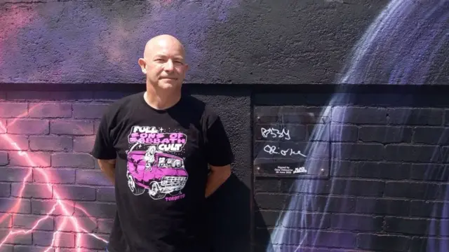 A bald man in a black and purple Black Sabbath top standing next to a wall mural that has been signed by Ozzy Osbourne. He is smiling and has his hands behind his back.