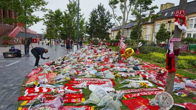 A man in black clothing crouches partially to take a picture of tributes to Diego Jota outside Anfield stadium in Liverpool