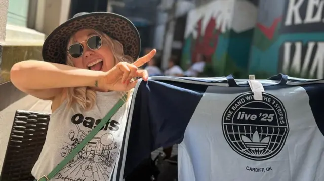 Sam Shiner posing with her tour merchandise in a sun hat, band t-shirt and shades