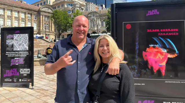 A bald man and a woman with blonde hair standing next to Black Sabbath advertising posters in a city centre square.