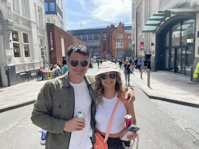 A man and a woman wearing sungalsses and bucket hats with the Principality stadium in the background. They are hoolding drinks cans. The man is wearing a green jacket and a white t-shirt. The woman is wearing a white vest top.