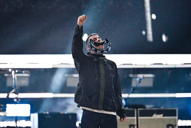 Liam Gallagher stands on stage at the Principality with a tambourine in his mouth. He holds one hand up in the air and wears a black parka jacket.