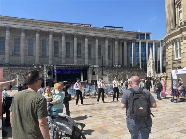 People walking around a city centre square where a food festival is taking place. There are three men playing brass instruments. The sky is blue and deck chairs can be seen in the background.