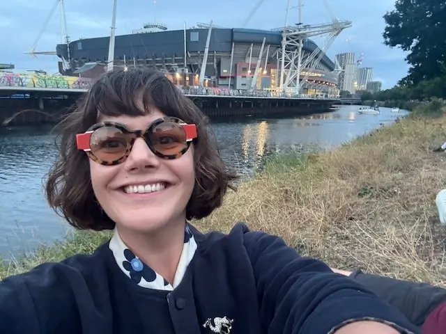 Georgina Rannard on the River Taff in Cardiff. She smiles at the camera and wears circular tortoiseshell glasses. She wears a navy bomber jacket and has brown short hair and a full fringe. The Principality stadium can be seen in the background.