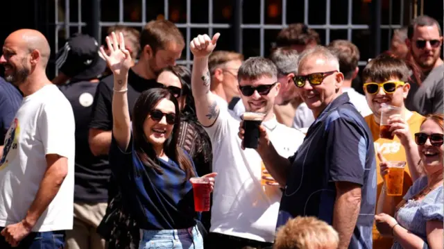 Crowds of people outside a pub holding drinks and chatting. A group of five, all wearing sunglasses are looking at the camera and smiling. They are all  holding pints and waving or giving thumbs up to the camera.