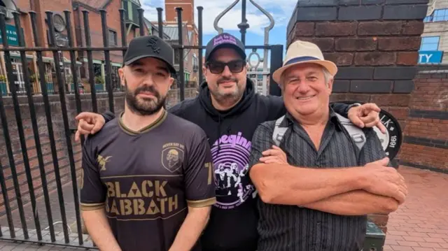 Three men across three generations standing in front of black railings. The man in the middle has his arms around the shoulders of the other two men and they are all smiling. Two of the men are wearing Black Sabbath t-shirts.