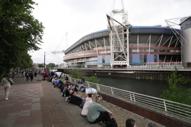 Rows of fans sit on the pavement along the River Taff in Cardiff, facing the Principality Stadium.