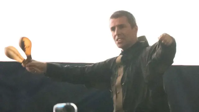 Liam Gallagher holds two maracas in his right hand, his left hand is also outstretched