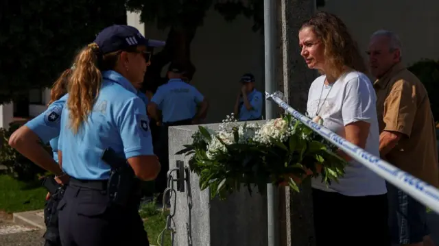 A woman in short-sleeved white shirts hands over a crown of flowers to a Portuguese police officer in blue cap, light blue polo shirt and black trousers