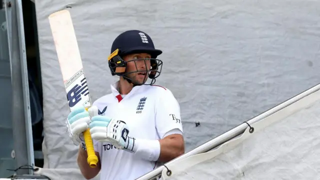 A man, Joe Root, holding a cricket bat with a cricket helmet on.