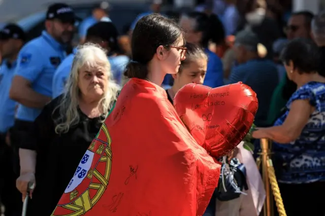 A young girl wearing a Portugese flag and holding a heart balloon for the queue to the public wake
