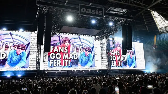 Oasis crowd stood in front of stage, big screen that says 'Fans go mad fer it!'