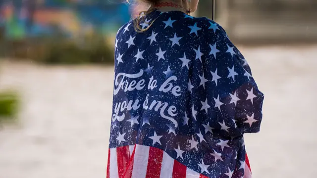 A woman with an American flag draped across her shoulders looks at flood water in Texas