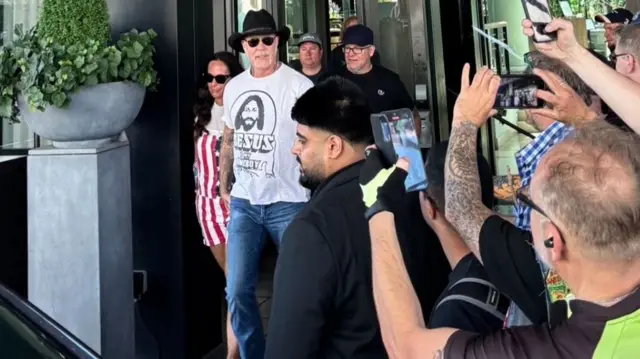 An older man, James Hetfield, wearing jeans and a white top, as well as a cowboy hat and sunglasses. He is walking out of a hotel entrance and fans stand to the side of him.