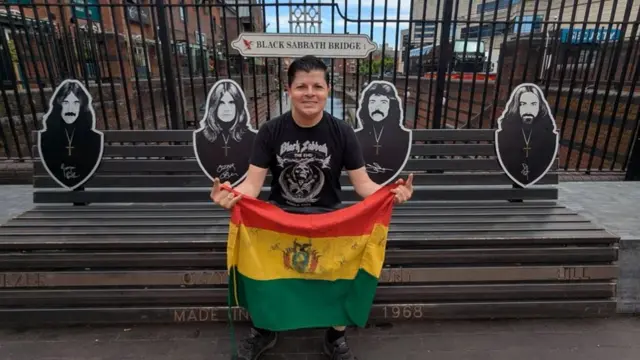 A man with short, dark hair sitting on a bench dedicated to Black Sabbath. He is holding a Bolivian flag.