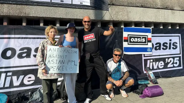 Four people standing on the pavement with Oasis live '25 branding behind them. In the middle is a girl with a blue bucket hat hoilding an anti-racism sign.