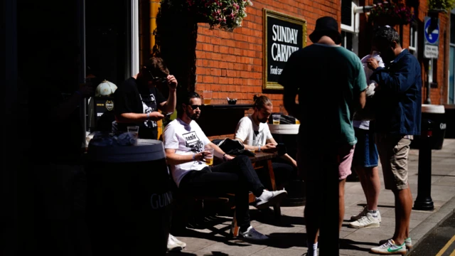A group of people sat at a wooden bench outside a pub, while others are stood on the pavement beside them. They are all shadinf their faces from the sun, and most are looking down at their phones. There are various pints in hands and on the table and one man is wearing a bucket hat.