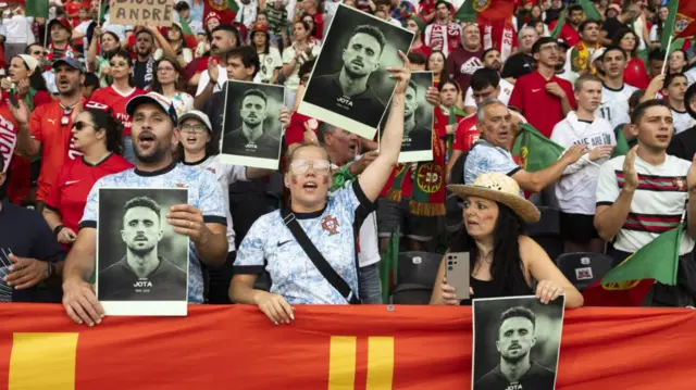 Portuguese fans hold up pictures of Diogo Jota inside a stadium, a young girl in glasses and two Portuguese flags painted on her cheeks at the front between a man and a woman. Behind them, several fans in Portugal red and green football kits and scarves