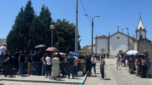 People stand outside a village in the heat, some with umbrellas. There is a media presence with cameras.